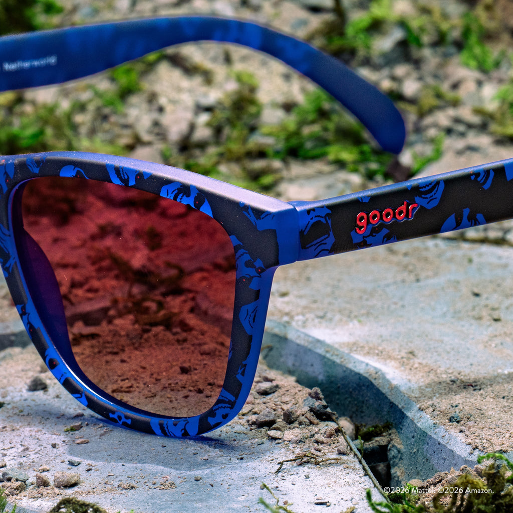 Close-up of blue and black Skeletor-patterned sunglasses with rose gradient lenses and goodr logo, set against a concrete background.
