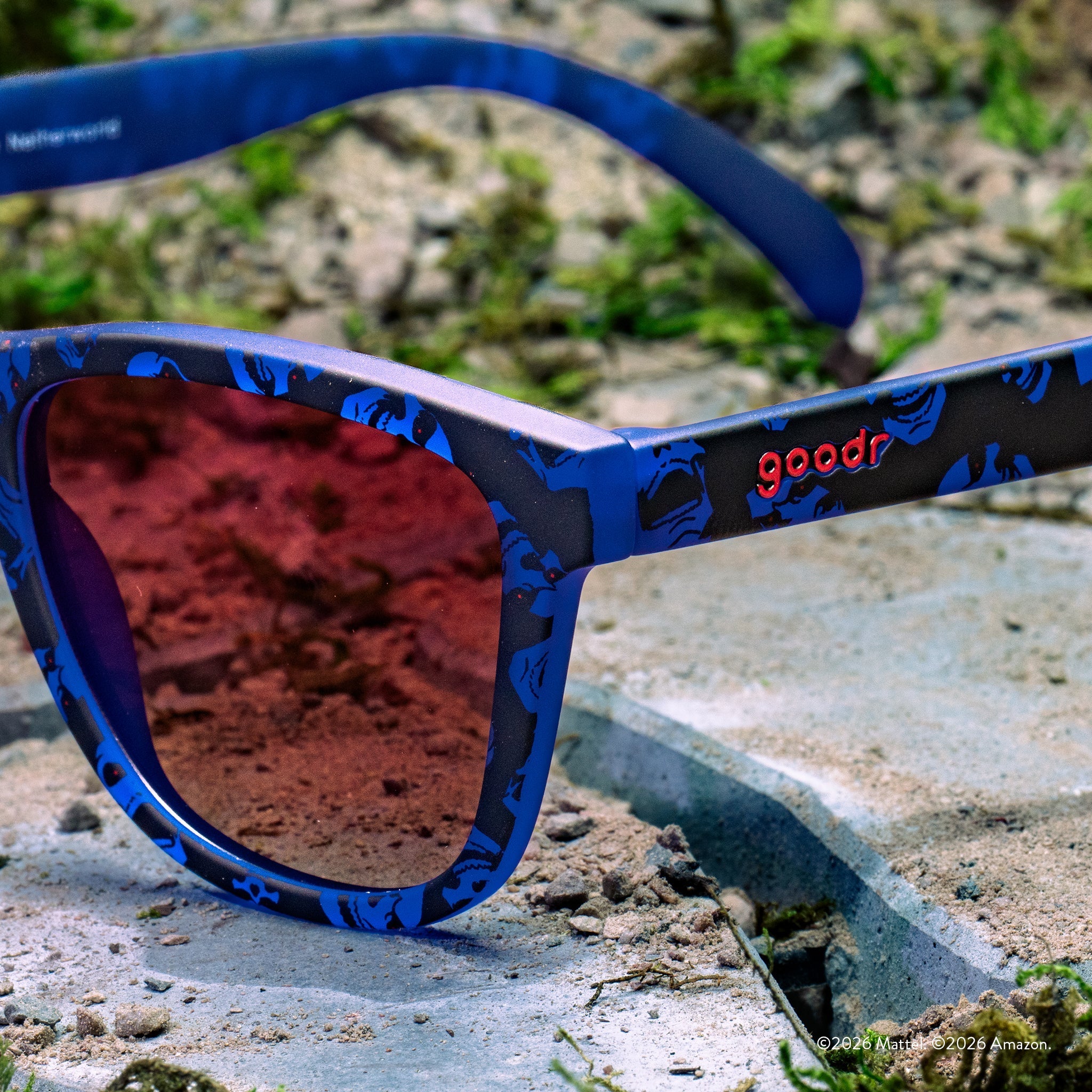 Close-up of blue and black Skeletor-patterned sunglasses with rose gradient lenses and goodr logo, set against a concrete background.