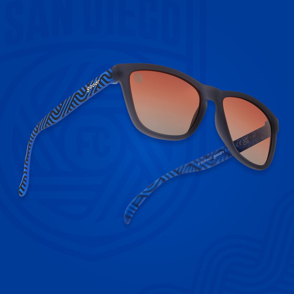 Angled view of shades tilted up. Dark gray frames w/ blue waves, San Diego FC on temples+orange gold gradient lenses.