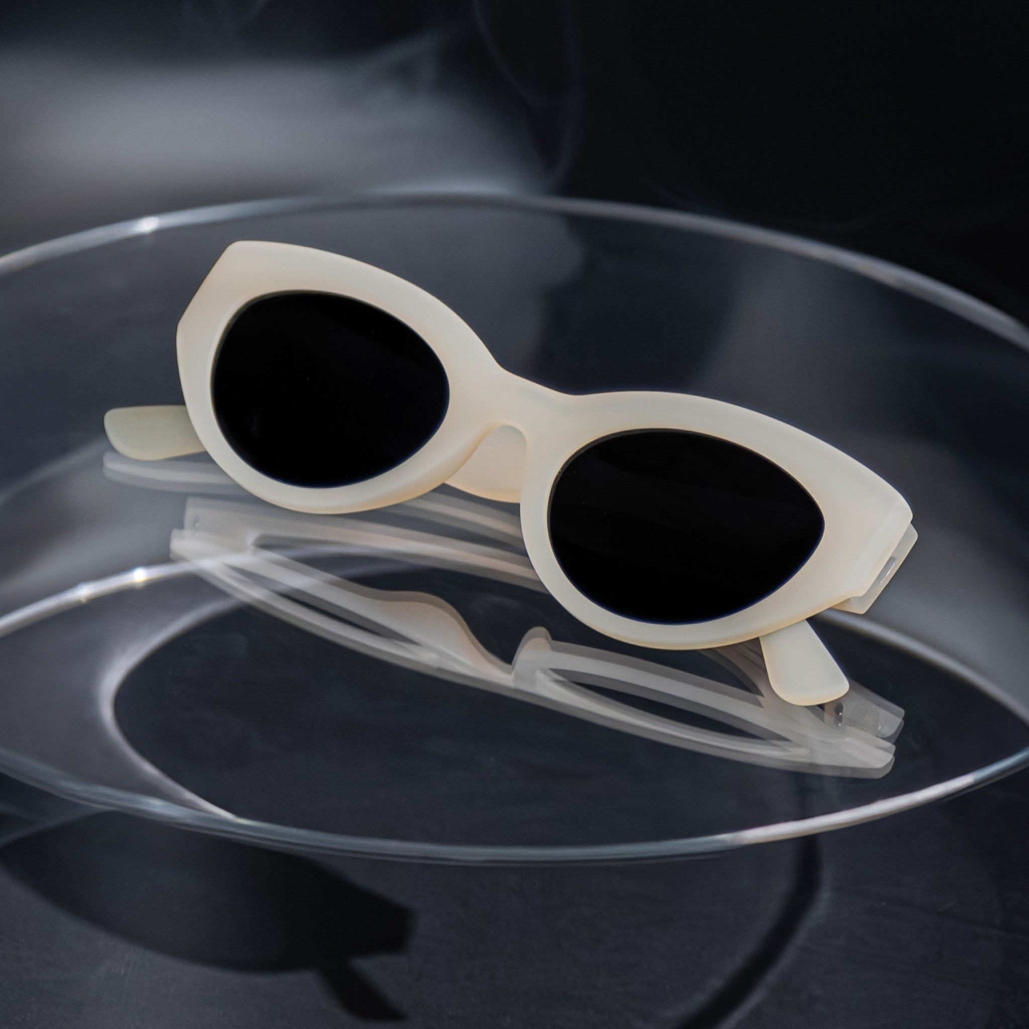 Close-up of translucent soft cream, retro-inspired angled cat-eye sunglasses with black non-reflective lenses, folded on a clear table.