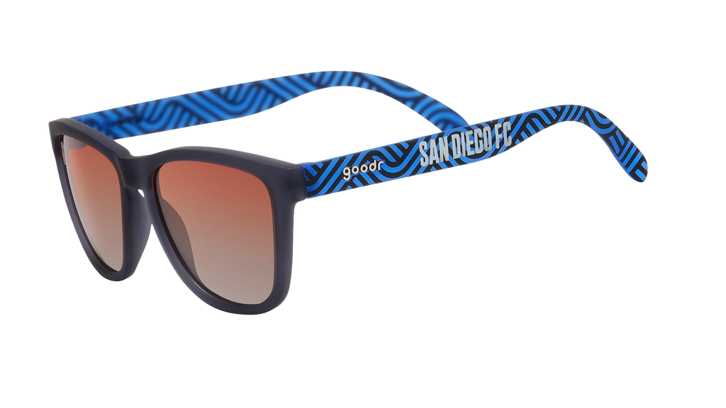 Angled view of shades tilted down. Dark gray frames w/ blue waves, San Diego FC on temples+orange gold gradient lenses.