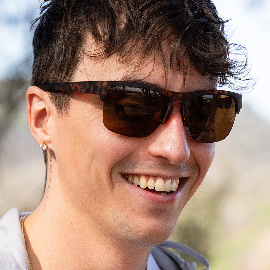 Close-up of a man outdoors, cheerfully smiling while wearing half-rim brown tortoiseshell sunglasses with brown non-reflective lenses.