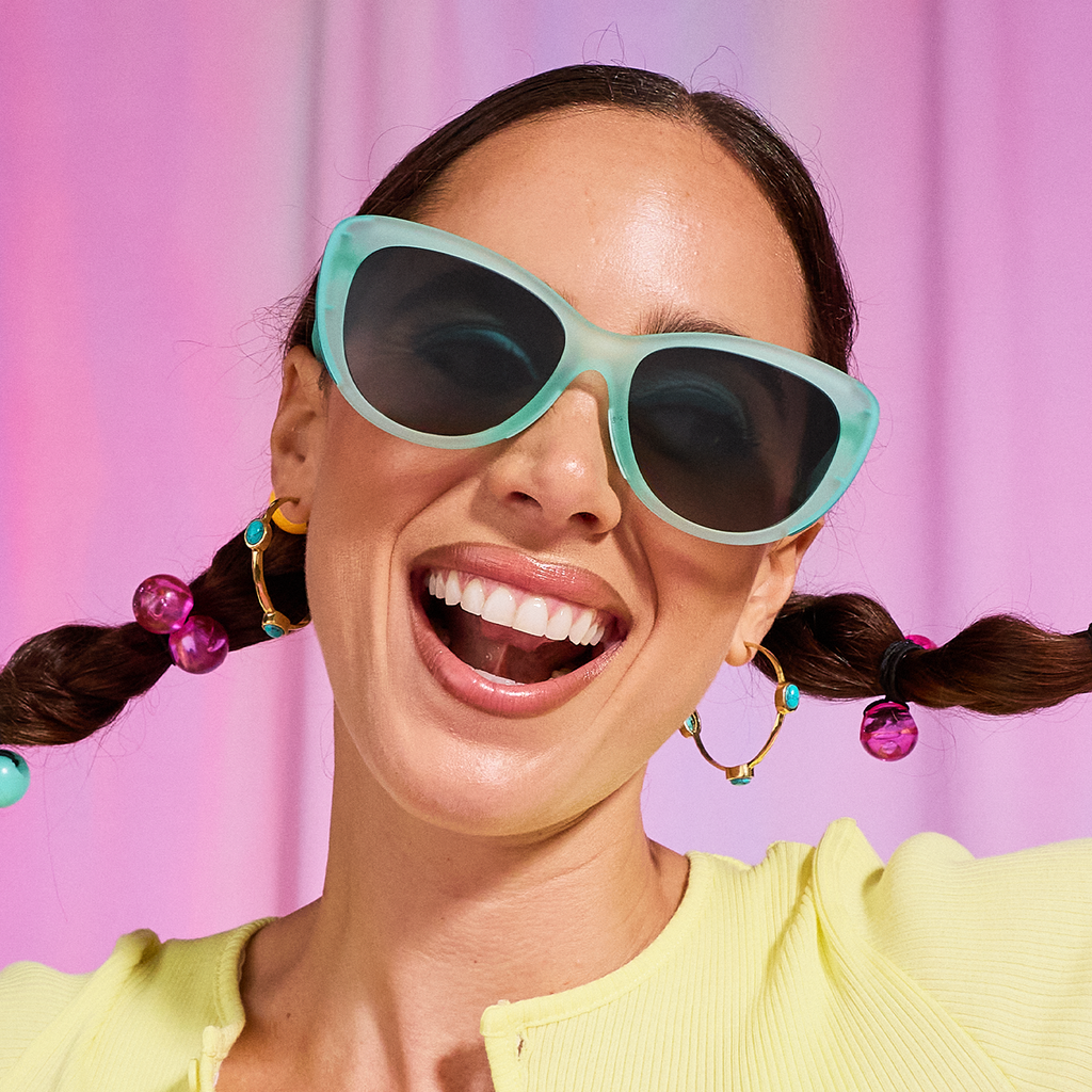 A woman with dark pigtails smiles excitedly as she models pale-green cat-eye sunglasses against a candy pink backdrop.