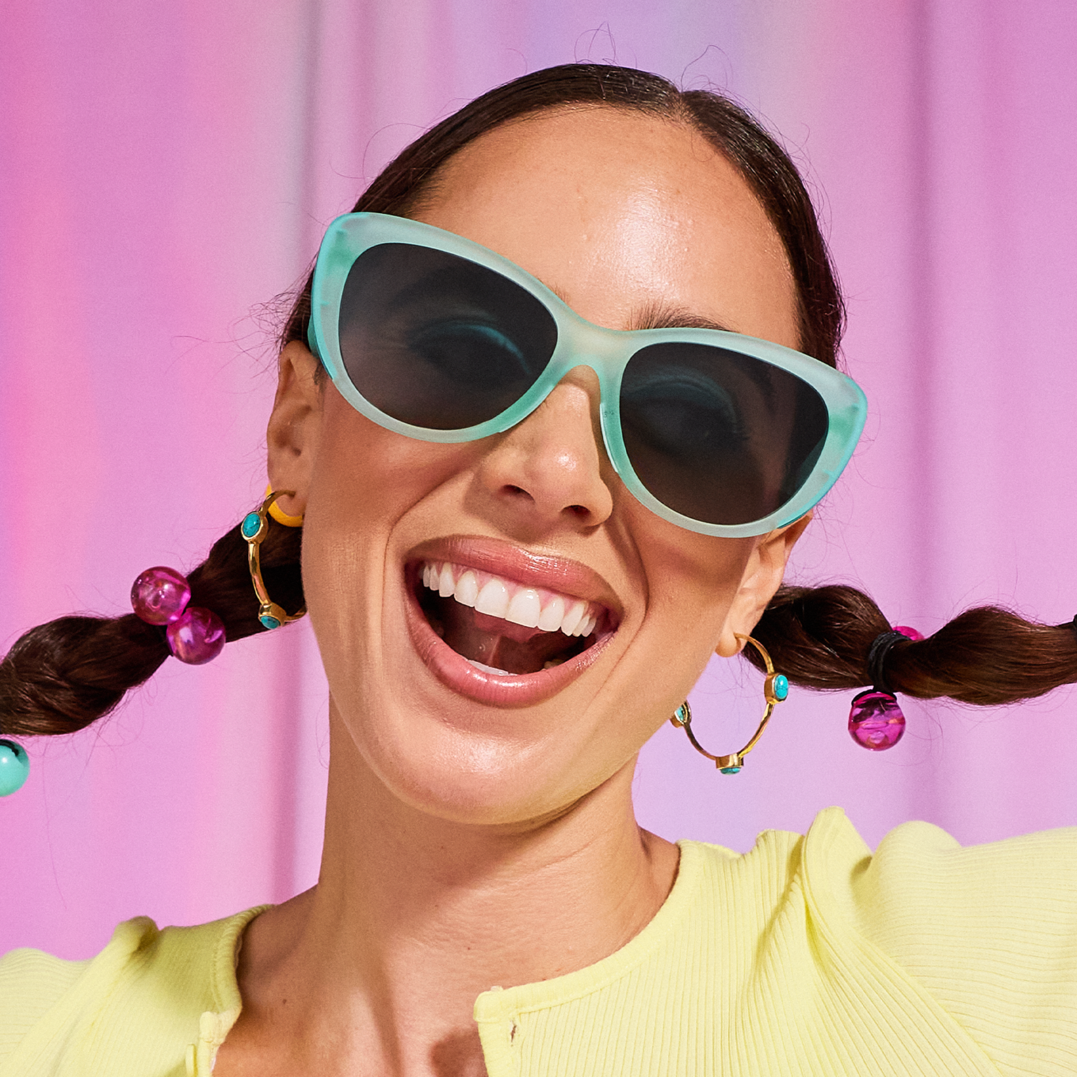 A woman with dark pigtails smiles excitedly as she models pale-green cat-eye sunglasses against a candy pink backdrop.
