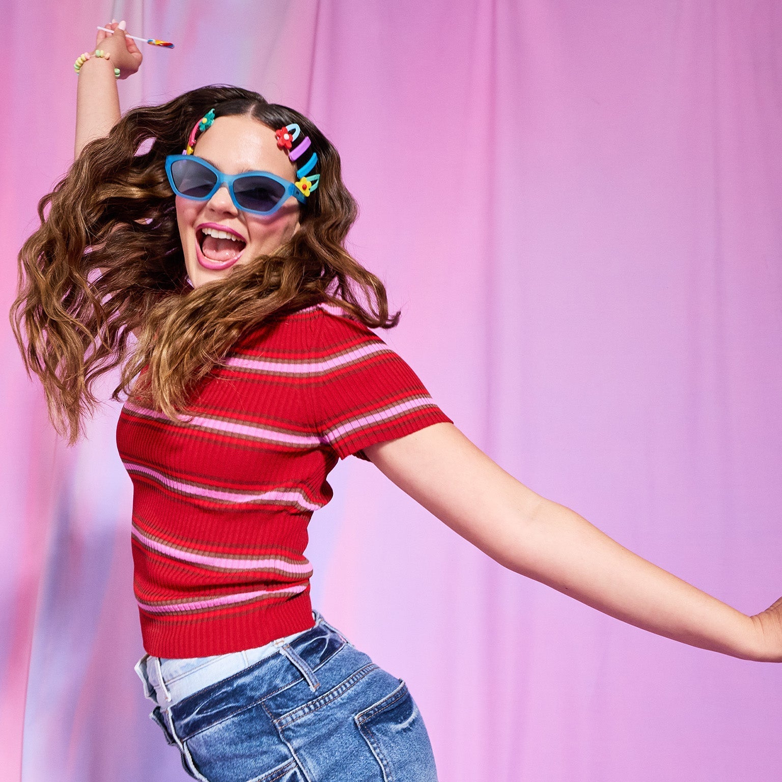 Young brunette woman in blue Deco G sunnies and colorful barettes jumping playfully, her arms outstretched and smile open.