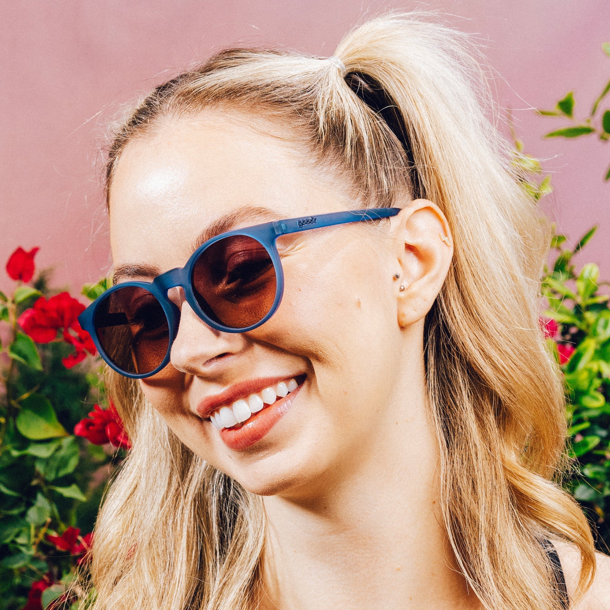 Woman smiling while wearing dark blue circular sunglasses with purple-yellow ocean lenses in front of a meadow backdrop.