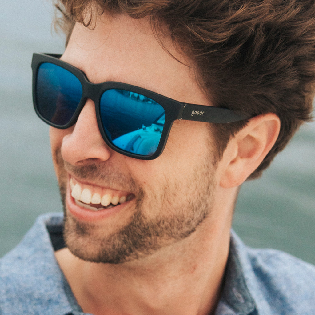 Three-quarter angle view of a happy young man with scruffy facial hair wearing black sunglasses with blue lenses.