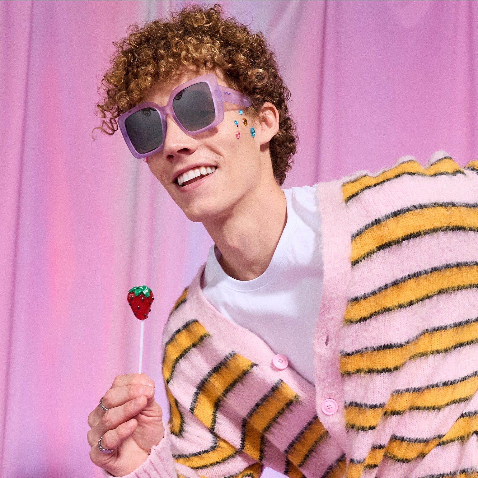 A young man with curly hair smiles while holding a lollipop that looks like a strawberry. He wears pale purple Amp G sunnies.
