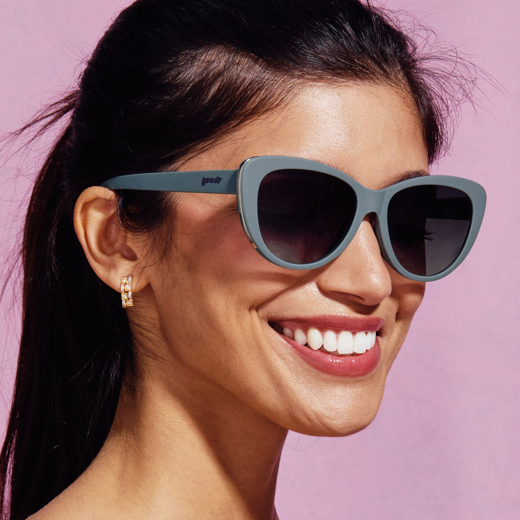 A woman smiling, wearing grey cat-eye sunglasses on a pink background.