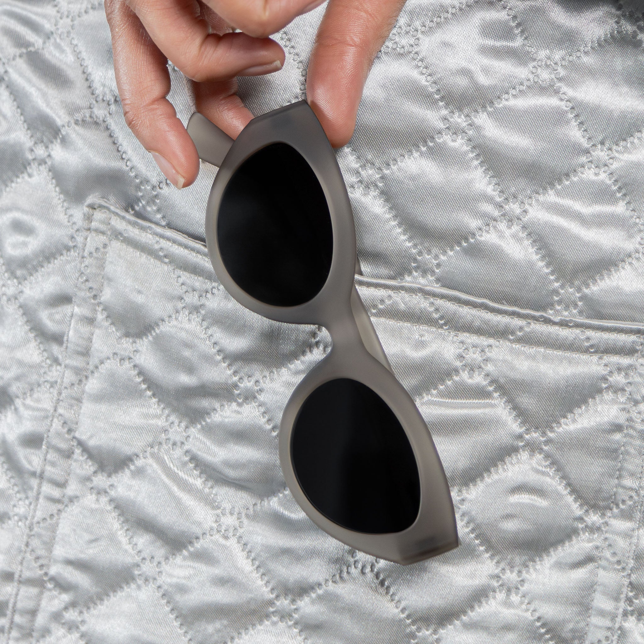 Close-up of a woman’s right side profile, wearing translucent dark gray, retro-inspired angled cat-eye sunglasses with black non-reflective lenses, folded on top of a light gray quilted fabric.
