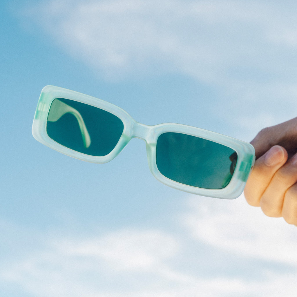 Masculine hand holding up translucent light green rectangular sunglasses with green lenses against the sky.