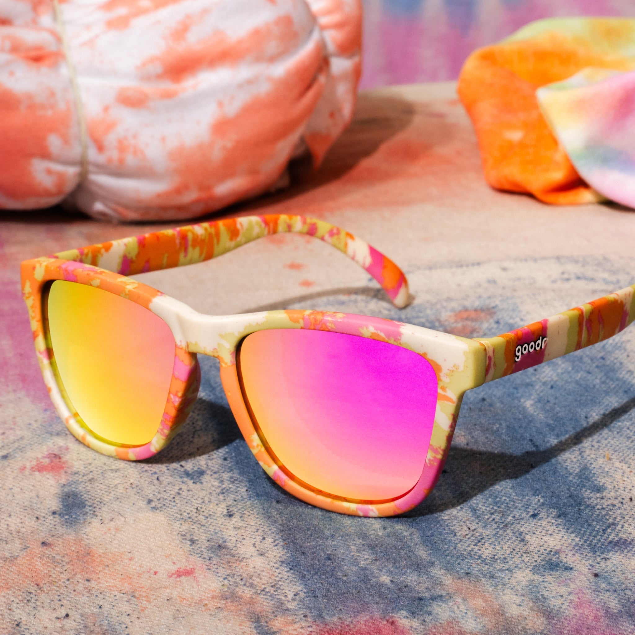 Three-quarter angle view of yellow, pink, and orange tie-dye shades with rose reflective lenses sitting on tie-dyed fabric.