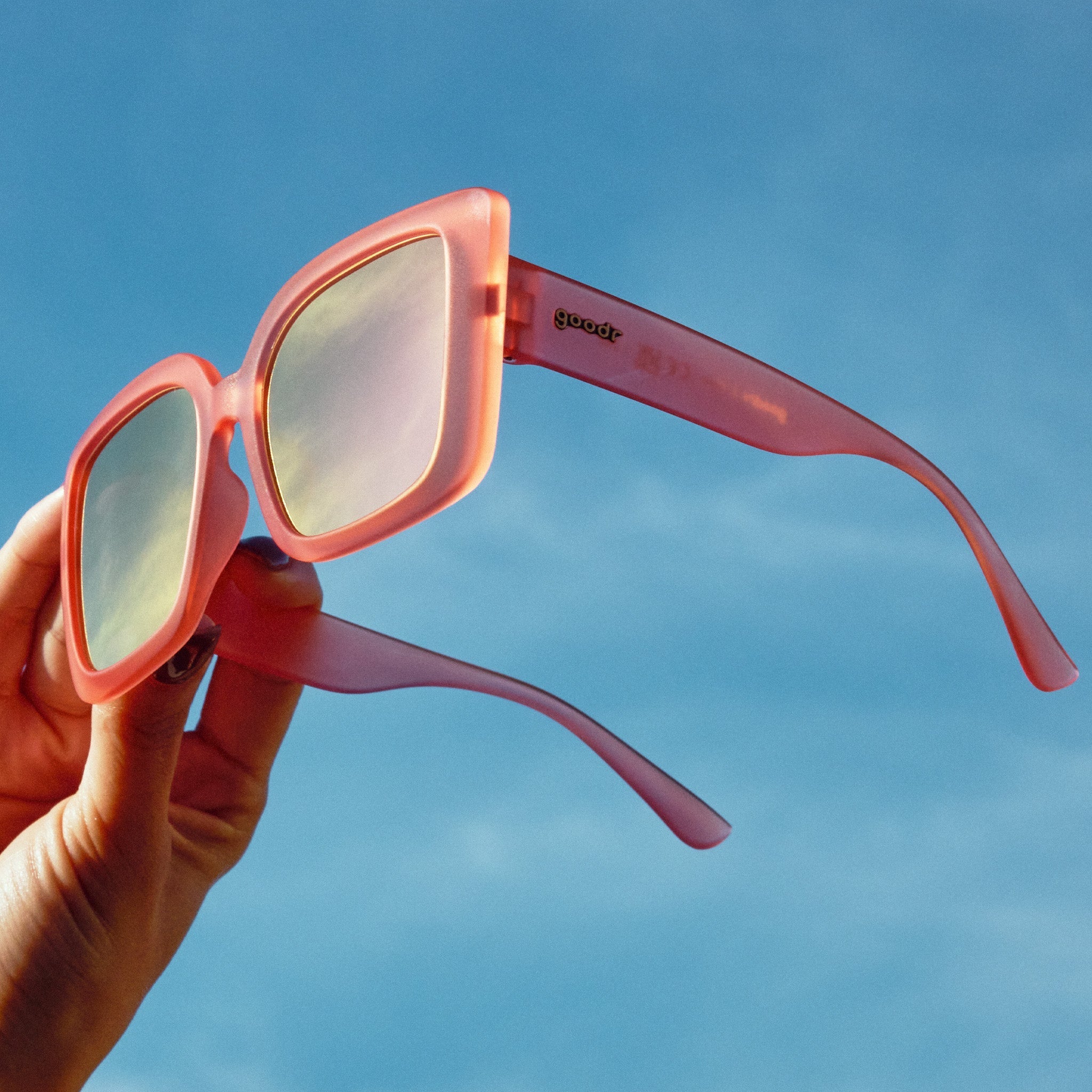 A hand holds a pair of oversized square sunglasses in coral pink against the sky at a three-quarter angle.