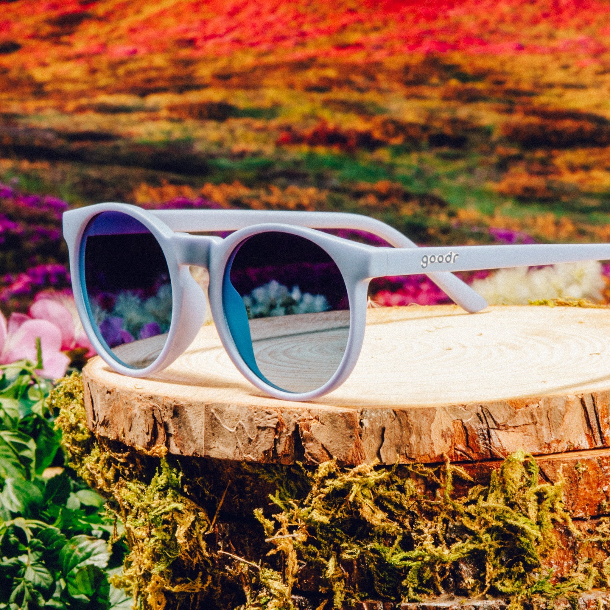 Three-quarter angle view of light blue circular sunglasses with dark blue gradient lenses against a meadow backdrop.