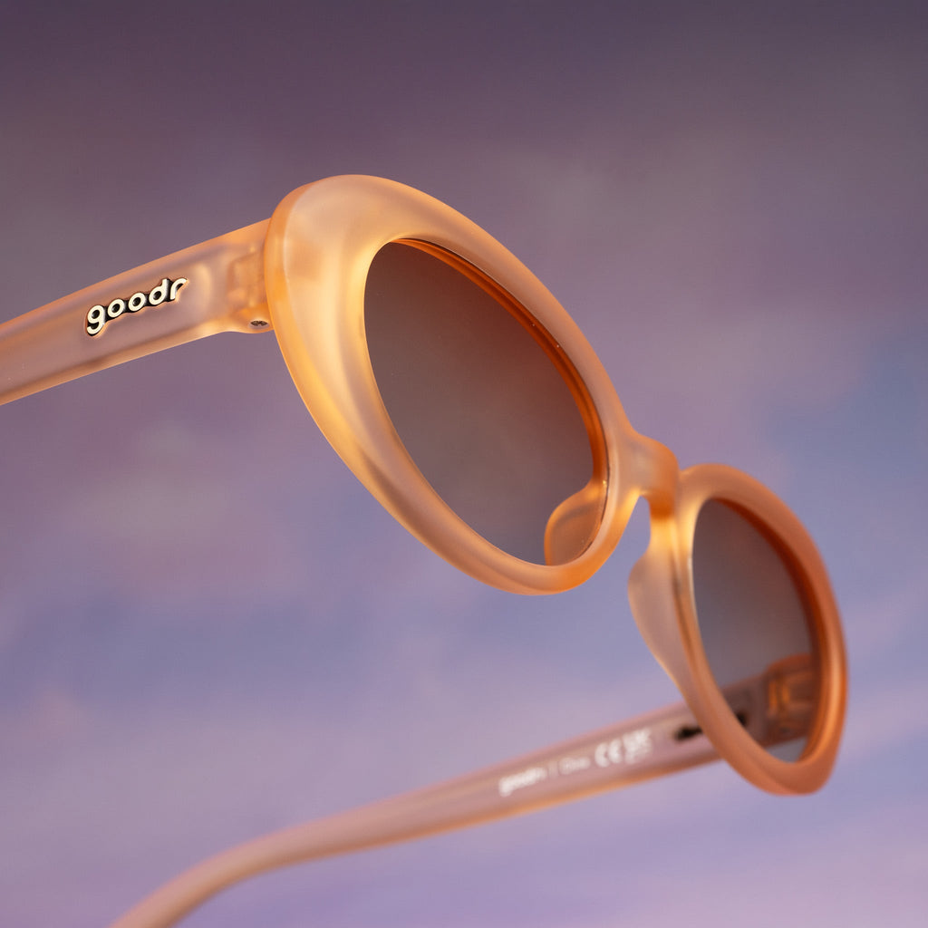 Three-quarter angle view of translucent pale peach oval Beat G sunglasses floating against a purplish-pink backdrop.