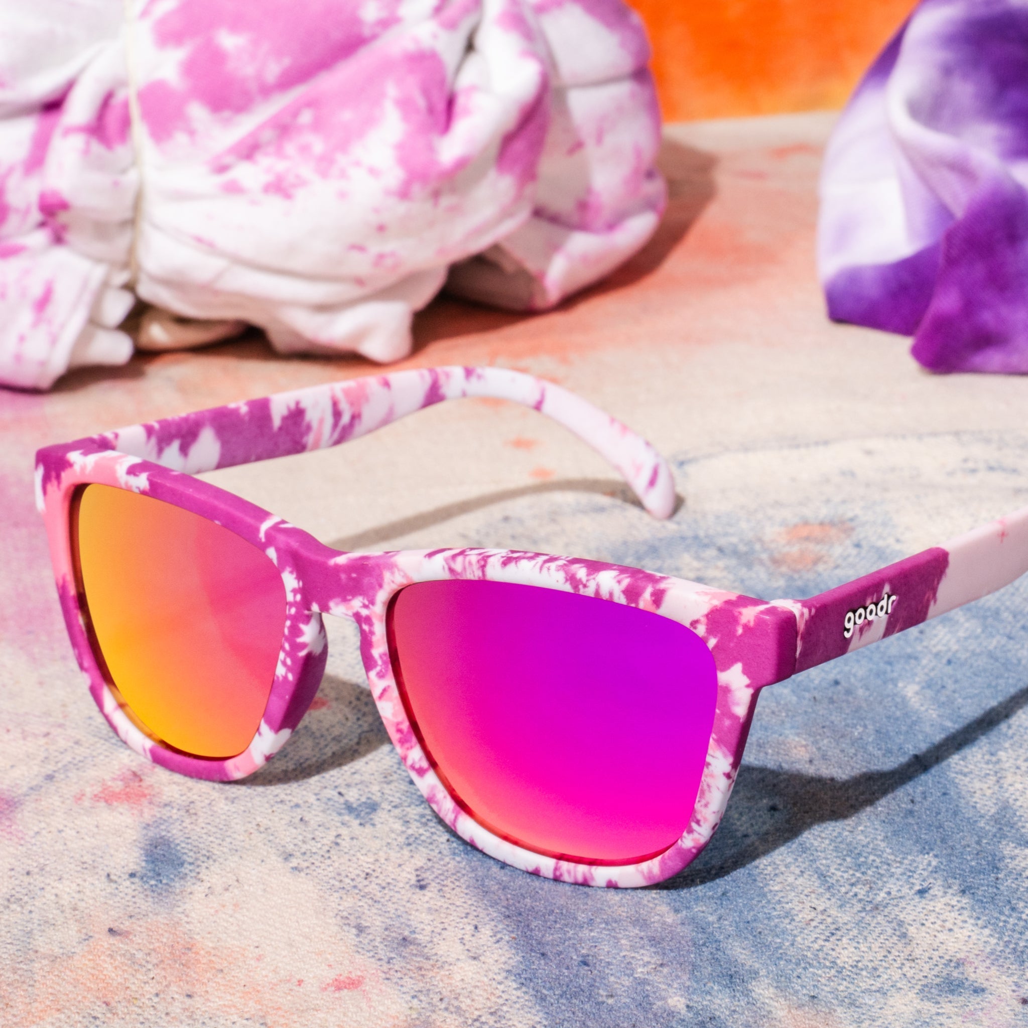 Three-quarter angle view of purple tie-dye sunglasses with rose reflective lenses sitting on a tabletop with tie-dyed fabric.