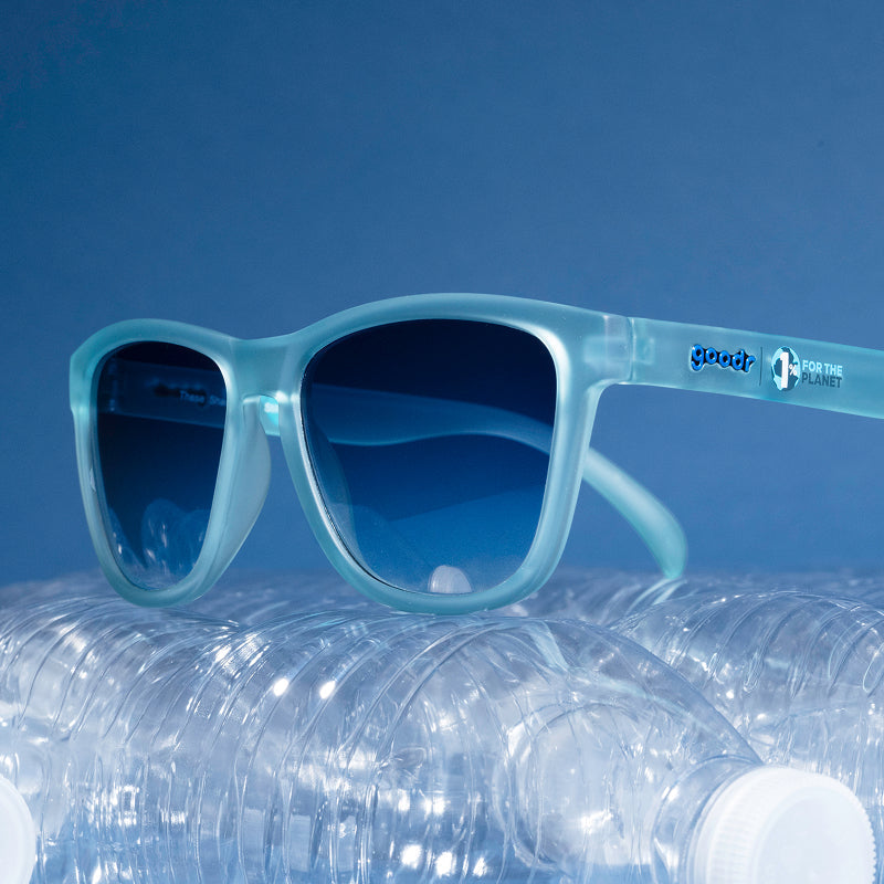 Open recycled paper box with filler holding translucent light blue sunglasses with blue lenses, bottles around.