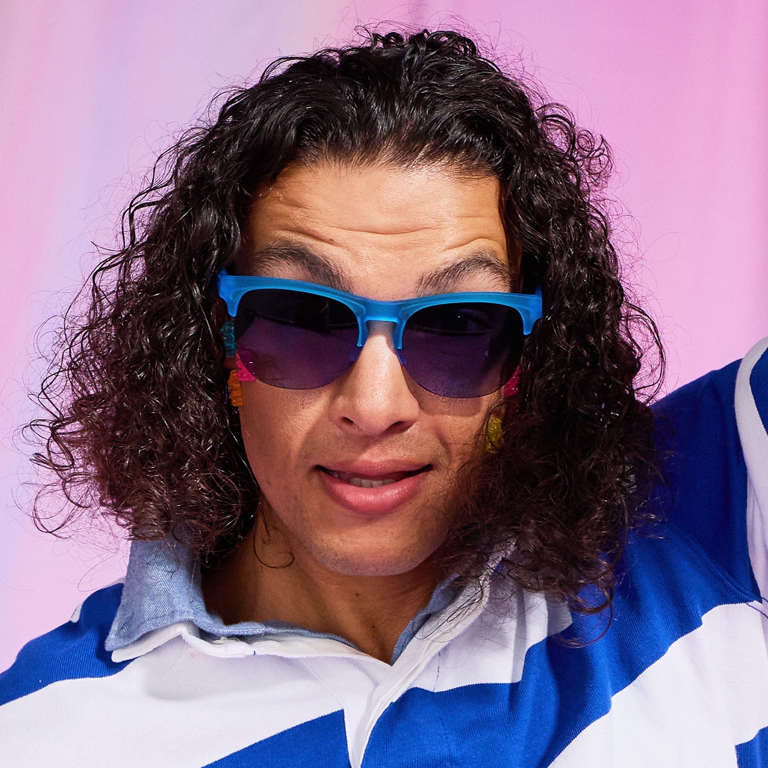 A man with dark curly hair and raised brows in a striped rugby shirt wears translucent blue half-rim Avant G sunglasses.