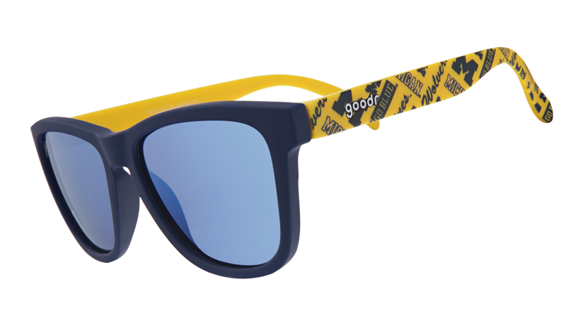 A pair of "goodr" sunglasses with navy-blue frames and reflective blue lenses. The arms of the sunglasses are bright yellow and feature University of Michigan "M" logos and text that reads "Go Blue" in a repeating pattern.