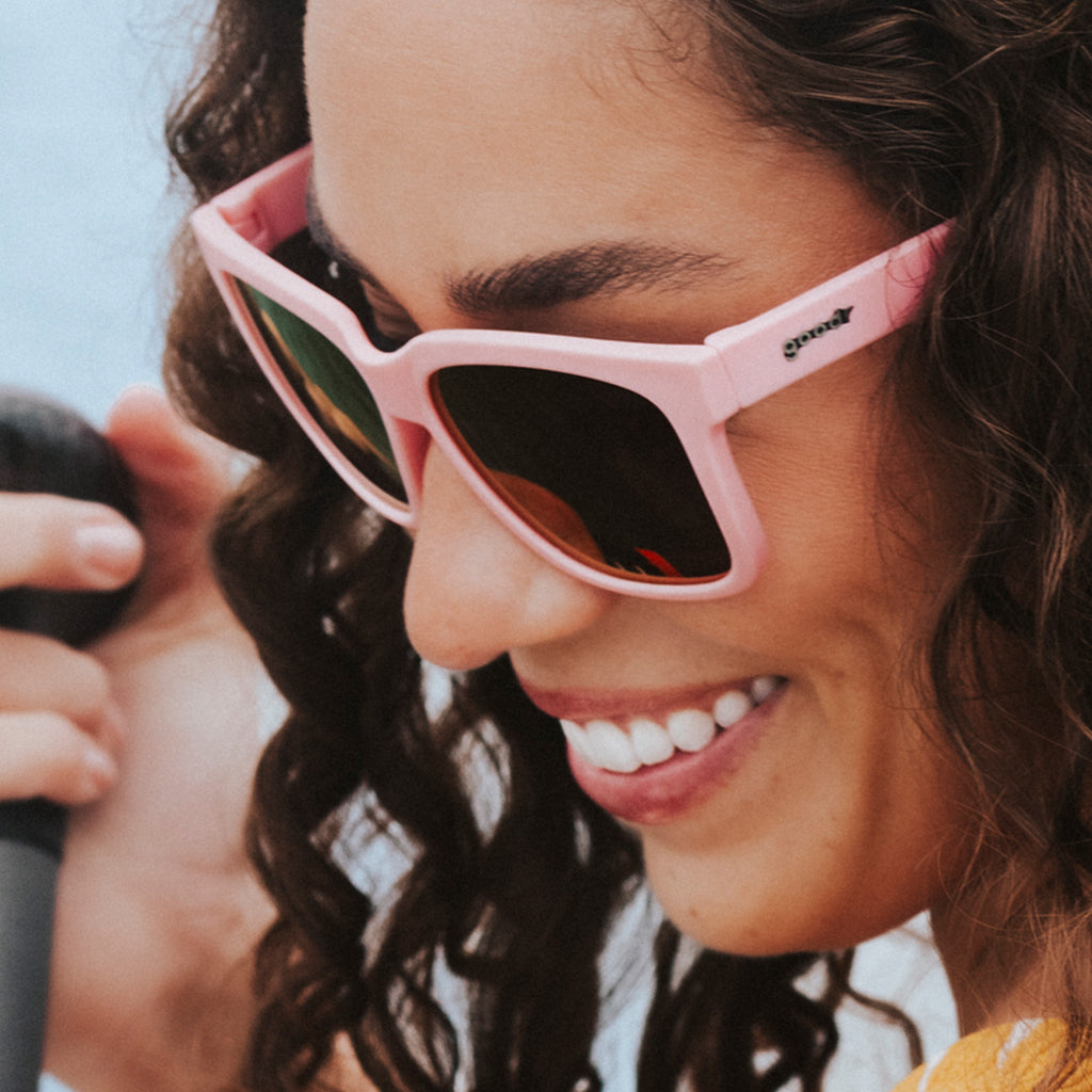 Side view of a smiling young woman with dark hair wearing pink sunglasses designed to float in water.
