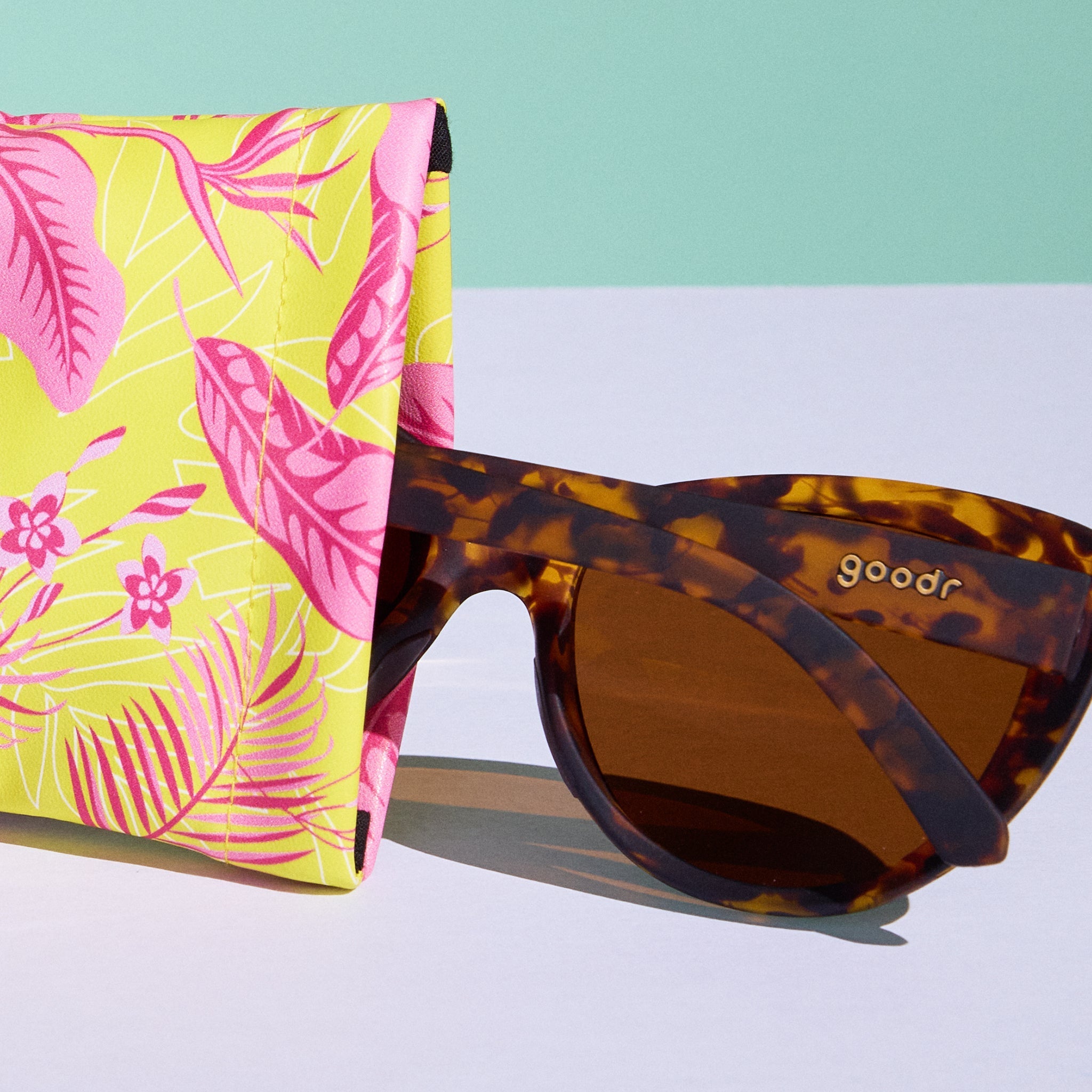 Close-up view of a soft fabric sunglasses case featuring a tropical design. A pair of tortoiseshell shades is shown partially pulled out.