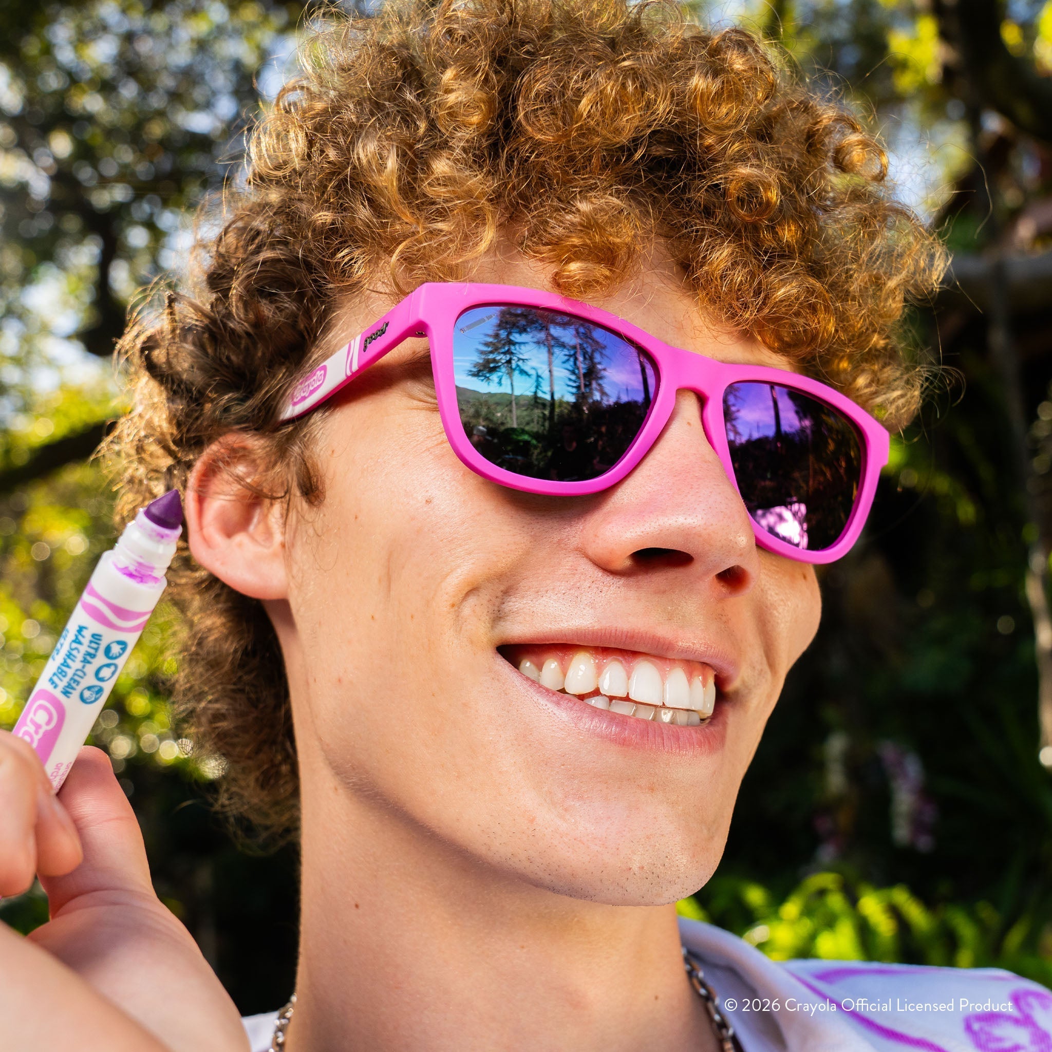 Man wearing magenta shades w/ white Crayola marker pattern+purple pink reflective lenses smiling in the outdoors.
