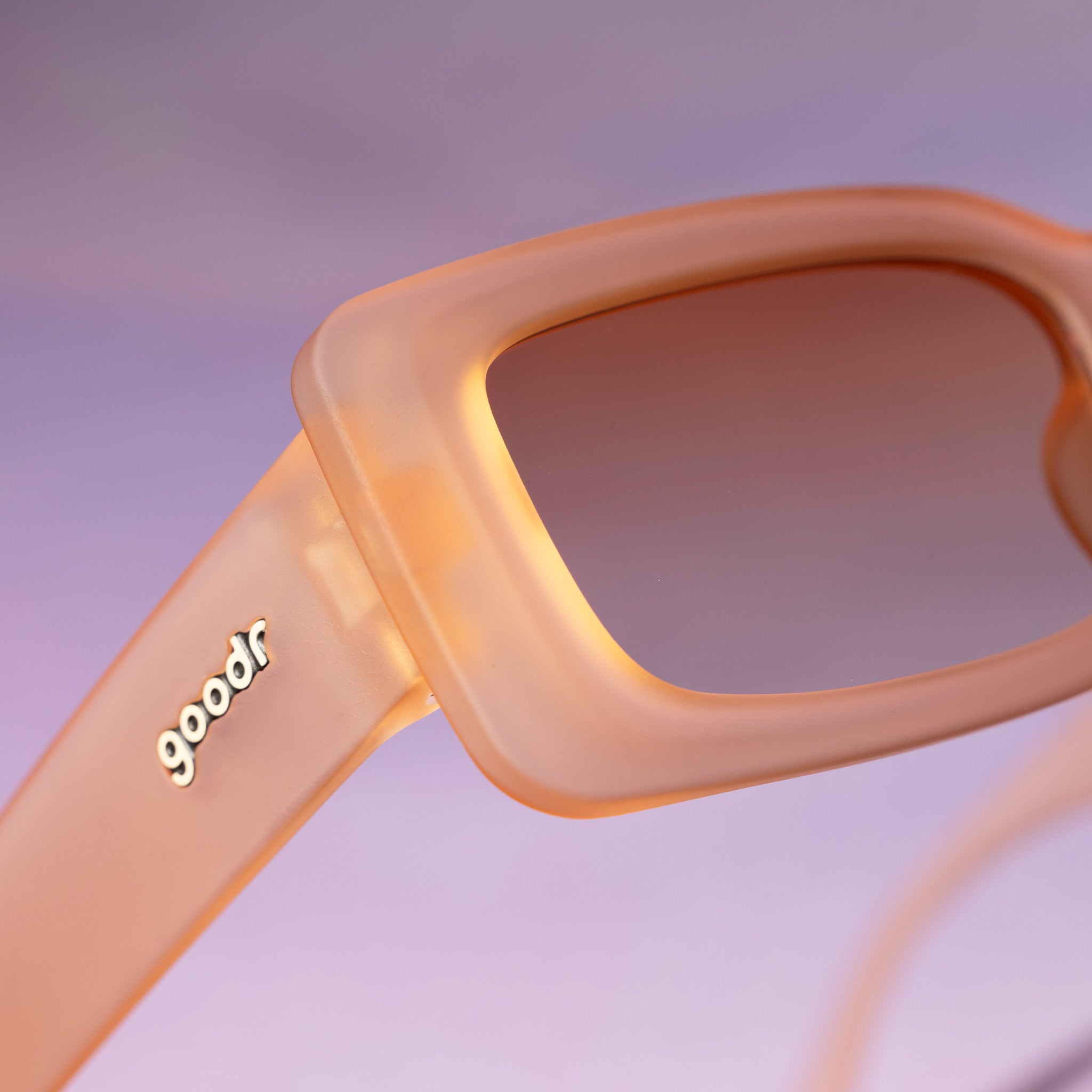 Close-up shot of the corner temple of a pair of translucent peach rectangular Luxe Gs against a purplish-pink backdrop.