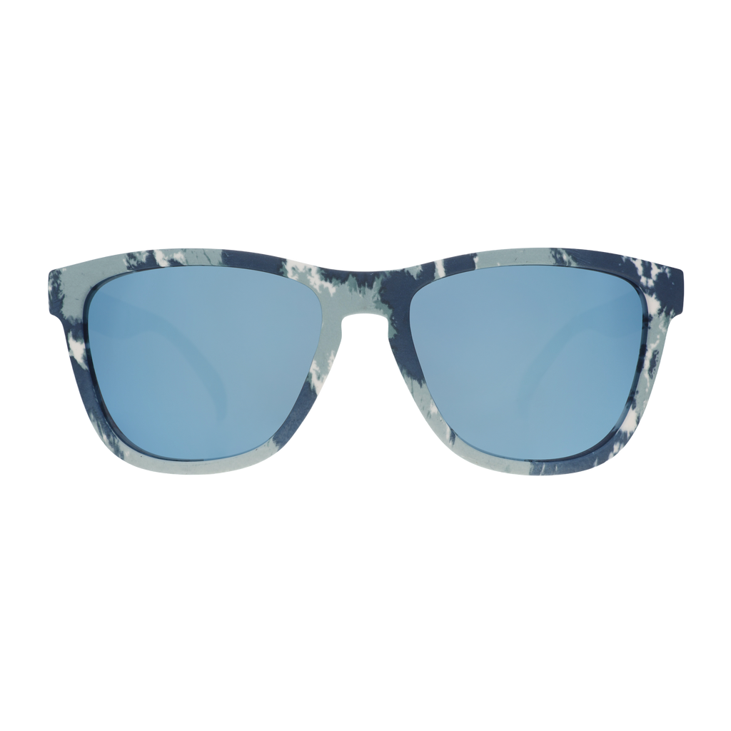 Three-quarter angle view of blue tie-dye sunglasses with blue reflective lenses sitting on a tabletop with tie-dye supplies.