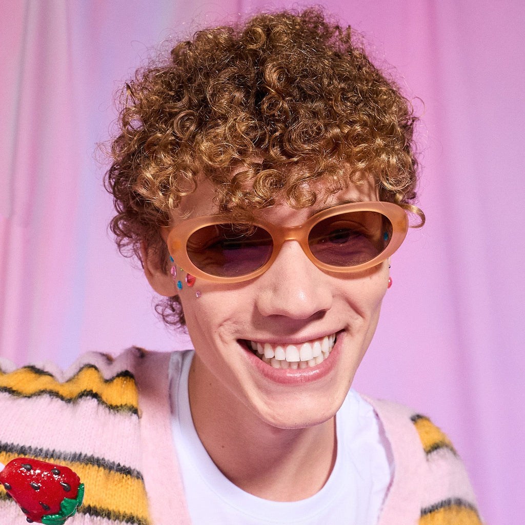A young man with strawberry blond curls smiles confidently in a pair of translucent pale peach oval Beat G sunglasses.