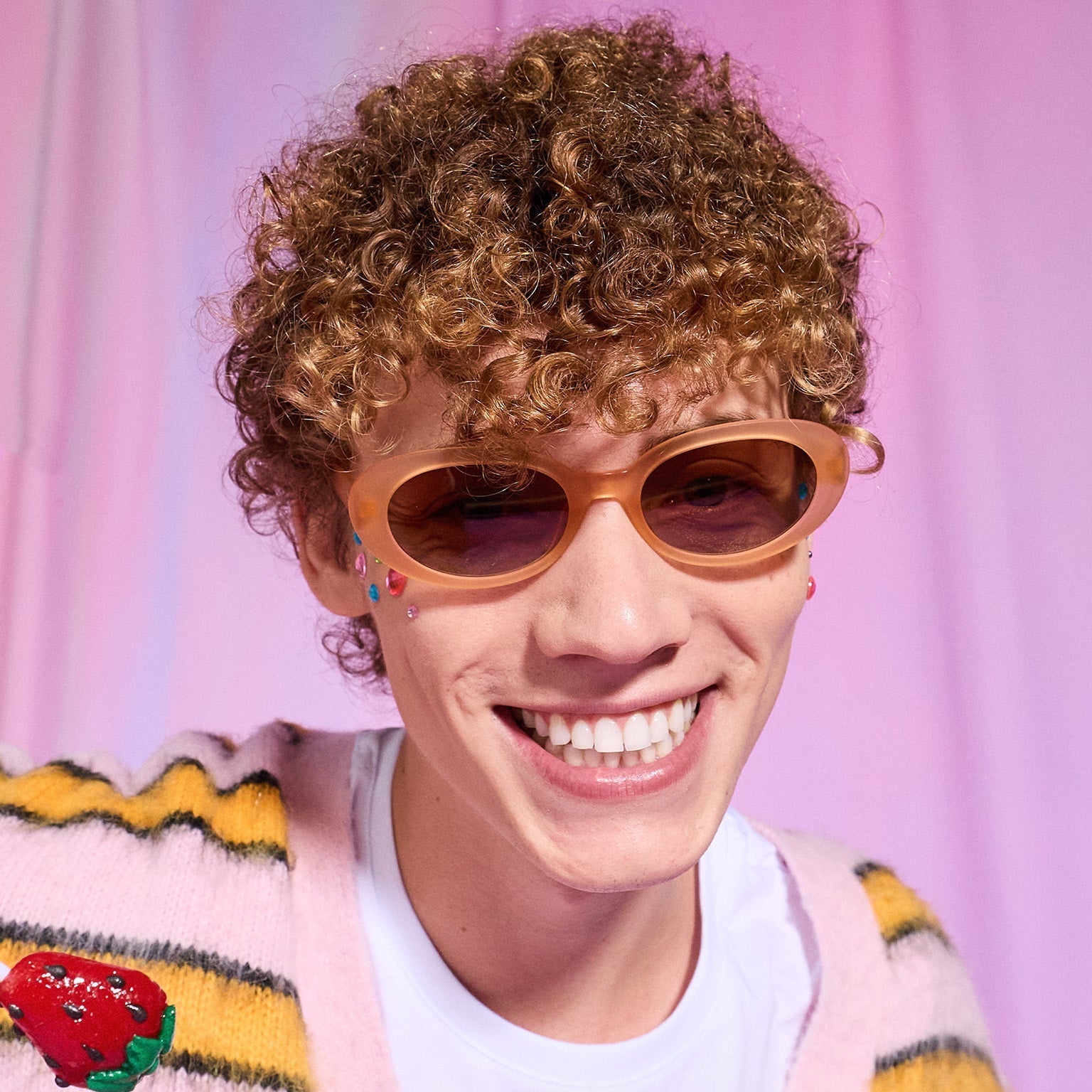 A young man with strawberry blond curls smiles confidently in a pair of translucent pale peach oval Beat G sunglasses.