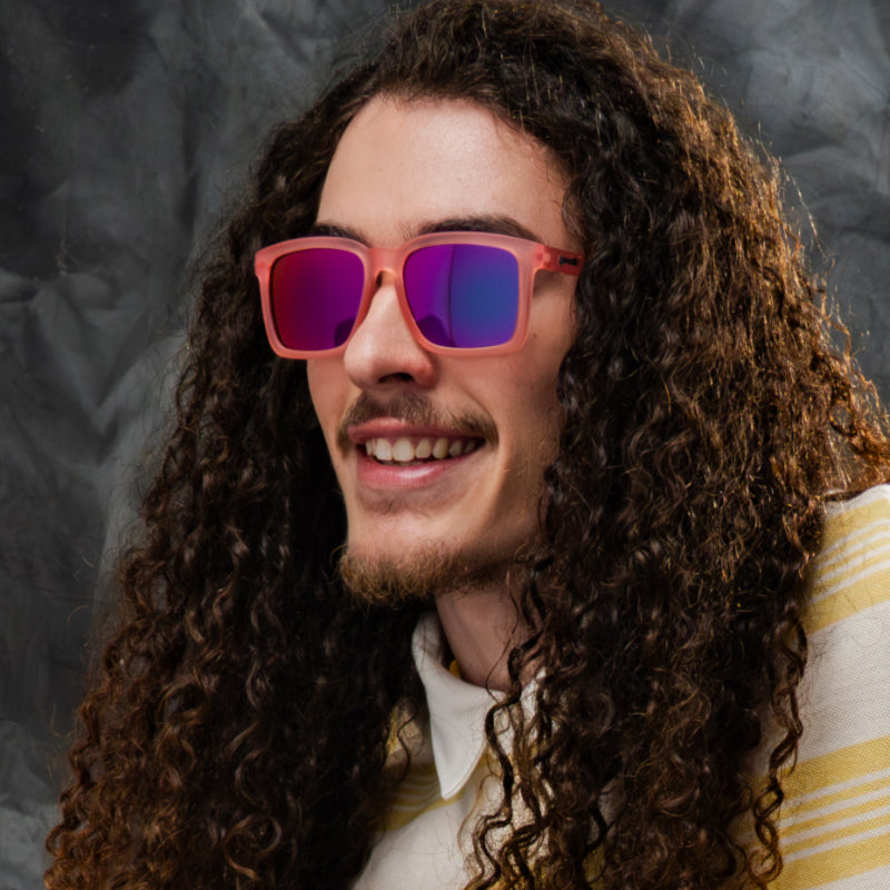 A man wearing translucent light pink shades with pink reflective lenses & a classic square silhouette is smiling.