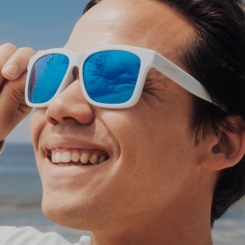 Side view of a smiling young man with dark hair wearing blue sunglasses designed to float in water.