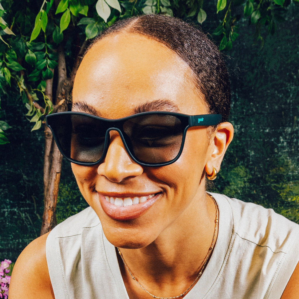 Woman smiling in front of a nature backdrop wearing translucent teal one-lens sunglasses with green gradient lenses.