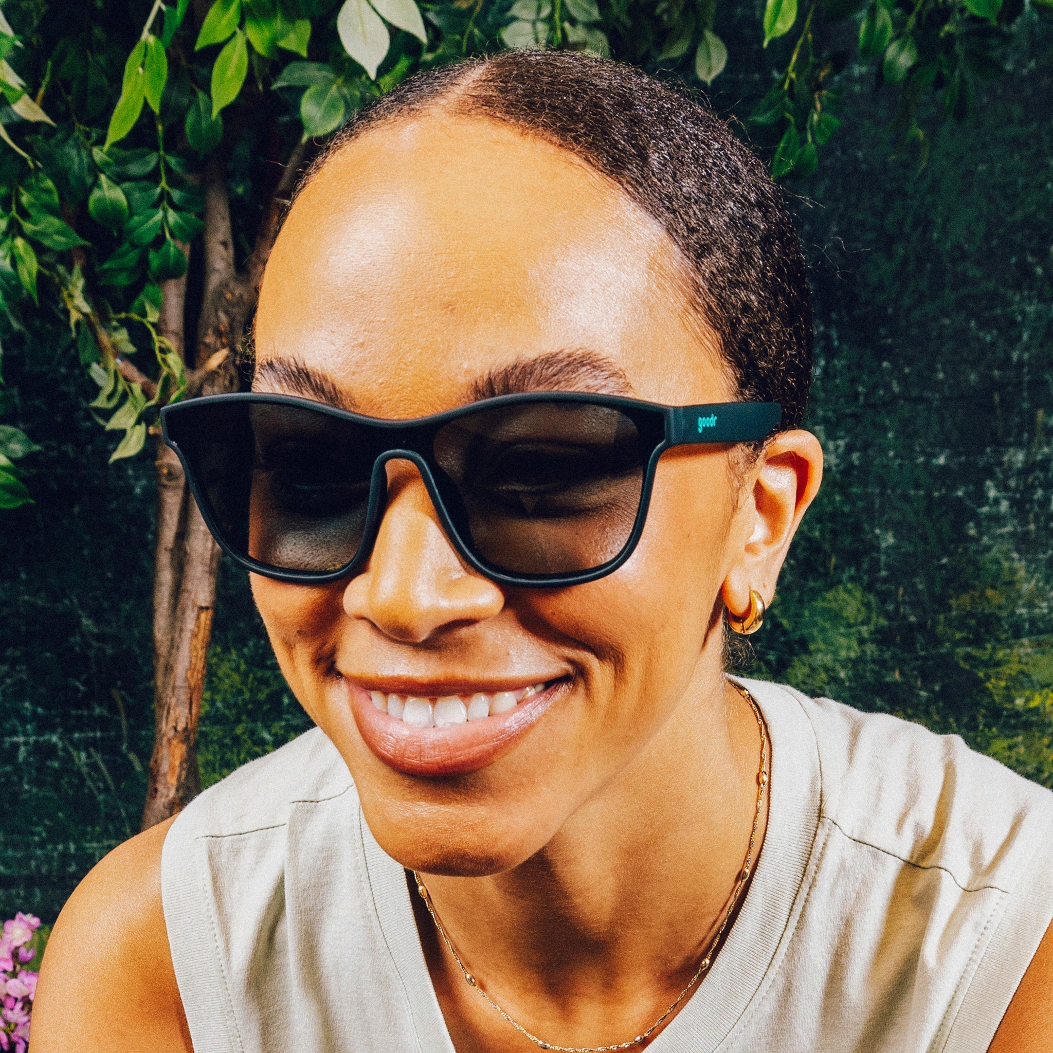 Woman smiling in front of a nature backdrop wearing translucent teal one-lens sunglasses with green gradient lenses.
