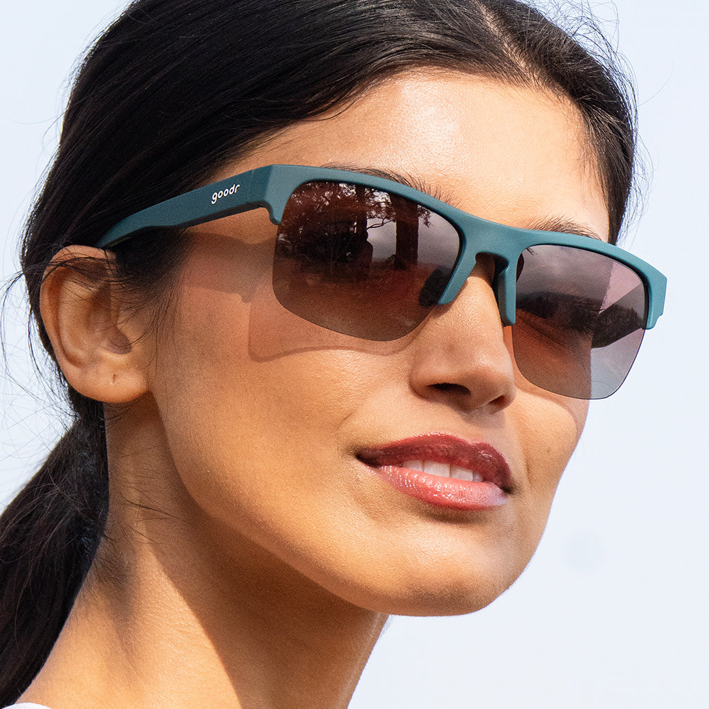 Close-up of a woman smiling while wearing solid green half-rim sunglasses with polarized rose gradient lenses.