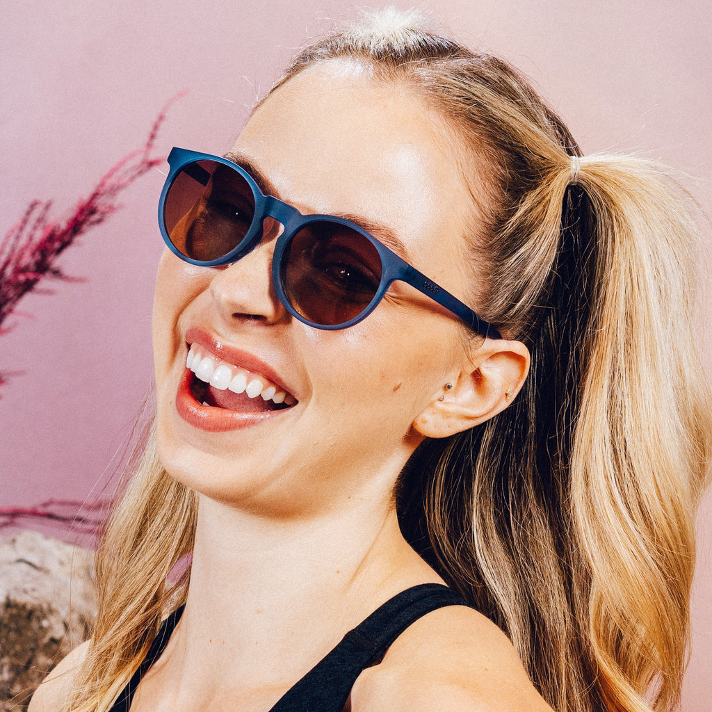 Side profile of a woman smiling and posing with dark blue circular sunglasses with purple-yellow ocean lenses.