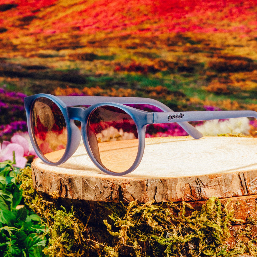 Three-quarter angle view of dark blue circular sunglasses with purple-yellow ocean lenses in front of a meadow backdrop.