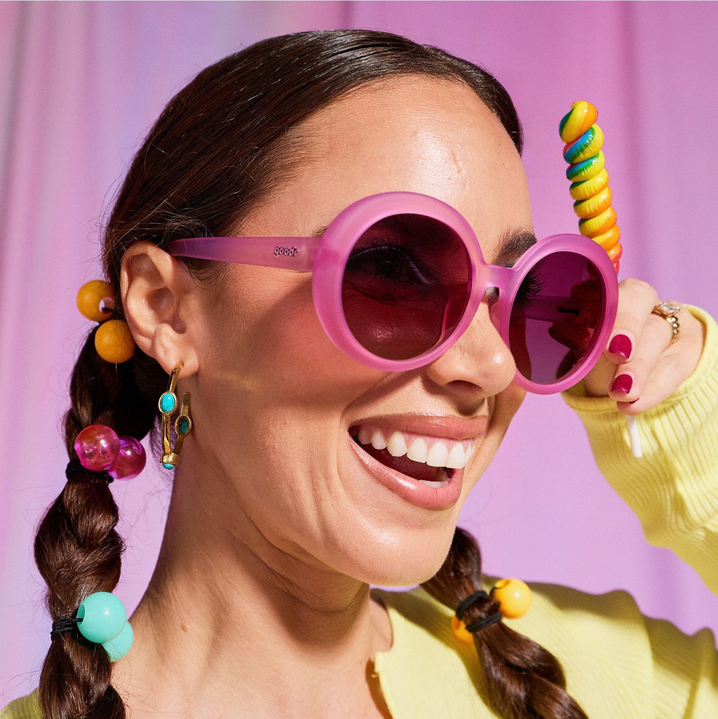 A woman with dark pigtails smiles confidently in a pair of translucent pink Mood G sunnies, holding a rainbow twirl lollipop.
