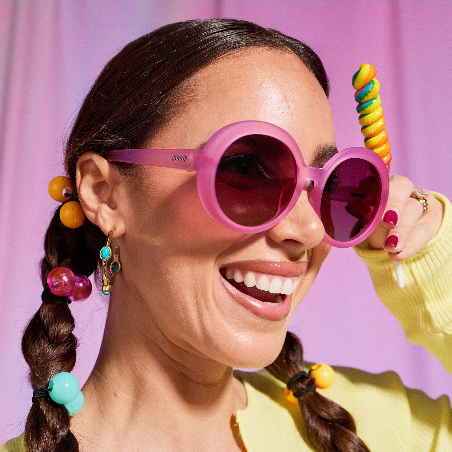 A woman with dark pigtails smiles confidently in a pair of translucent pink Mood G sunnies, holding a rainbow twirl lollipop.