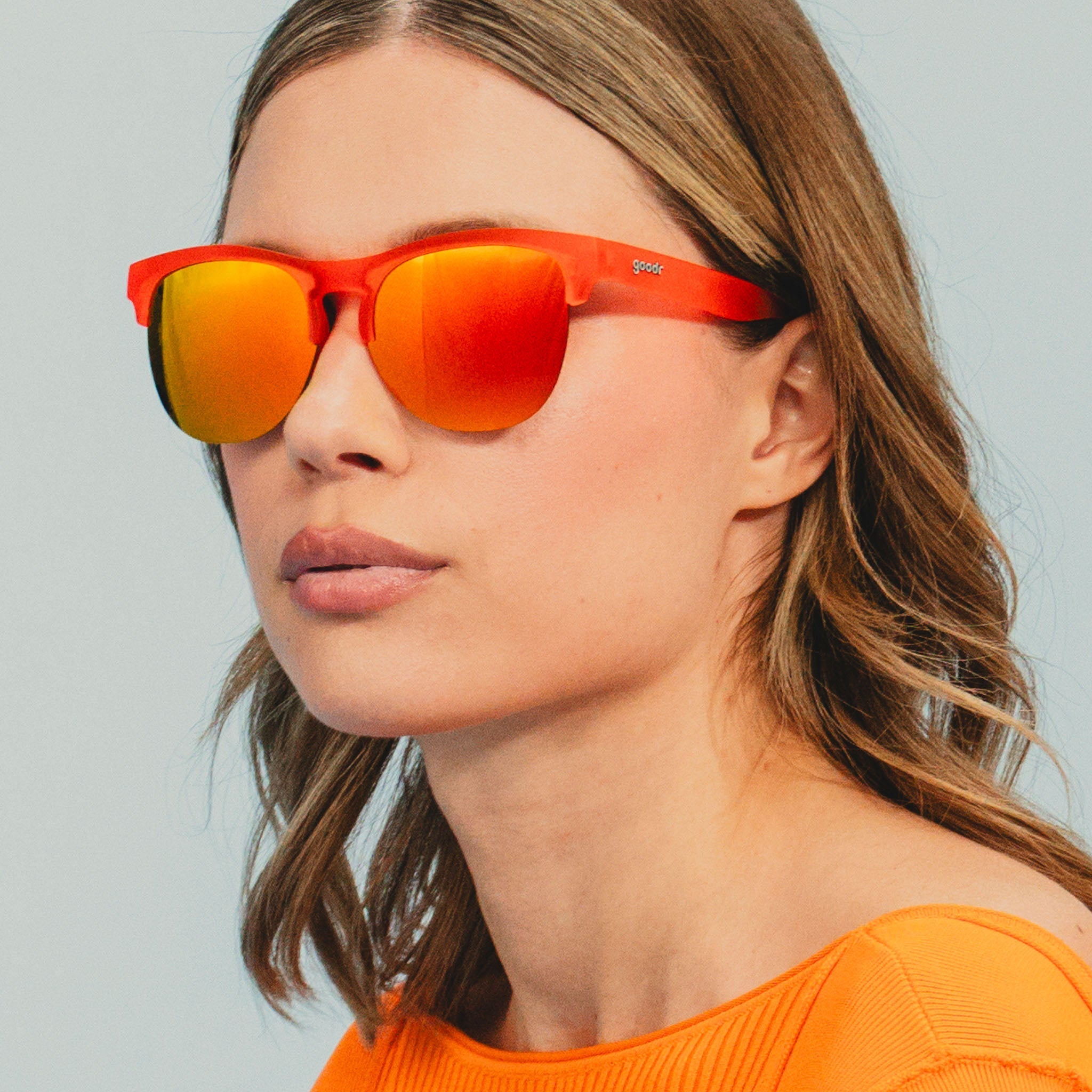 A smiling woman wears vibrant red-orange half-rim sunglasses.