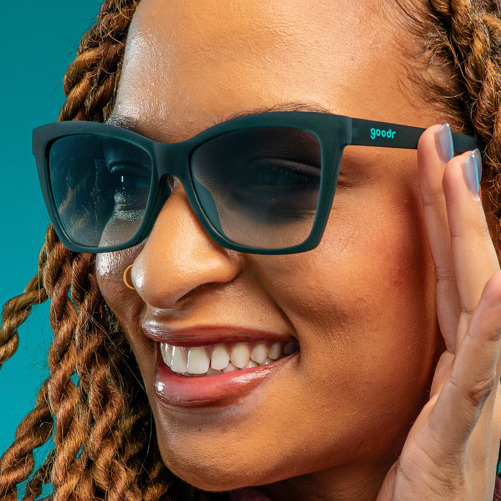 A woman with copper twist braids and a nose ring smiles in a pair of translucent teal Pop Gs with green gradient lenses against a teal backdrop.