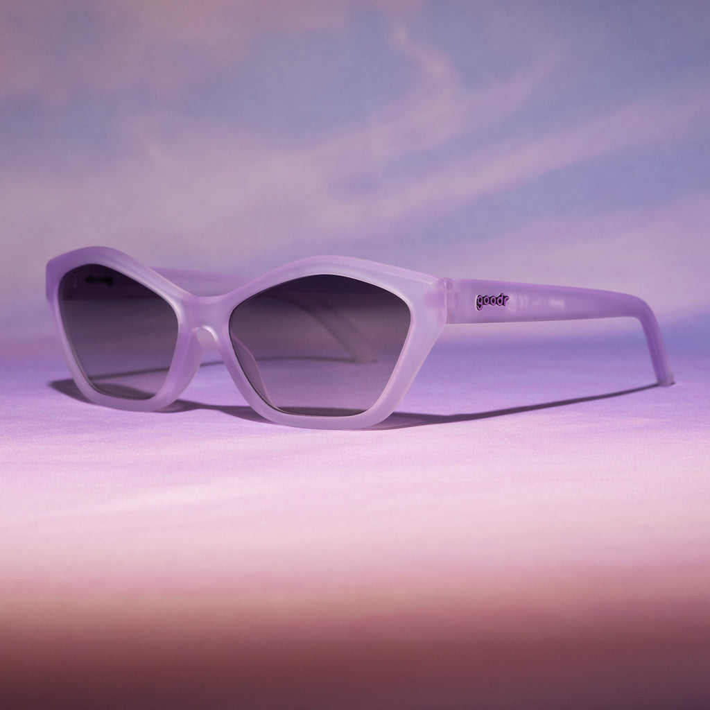 Three-quarter view of translucent pale purple Deco G sunglasses sitting on a purplish pink sunset-like backdrop.