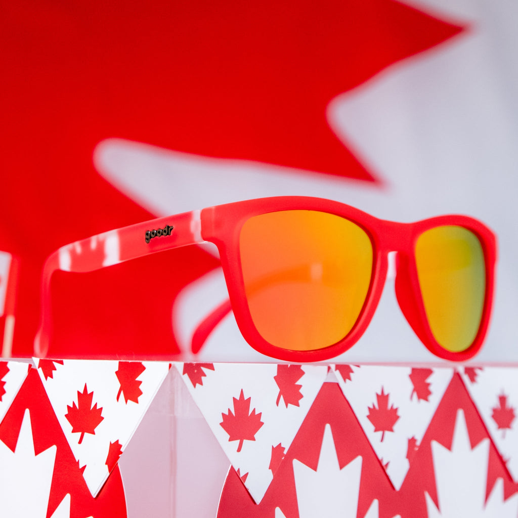 Angled view of red shades w/ a Canadian flag pattern+burnt orange reflective lenses on a podium w/ a maple leaf pattern.