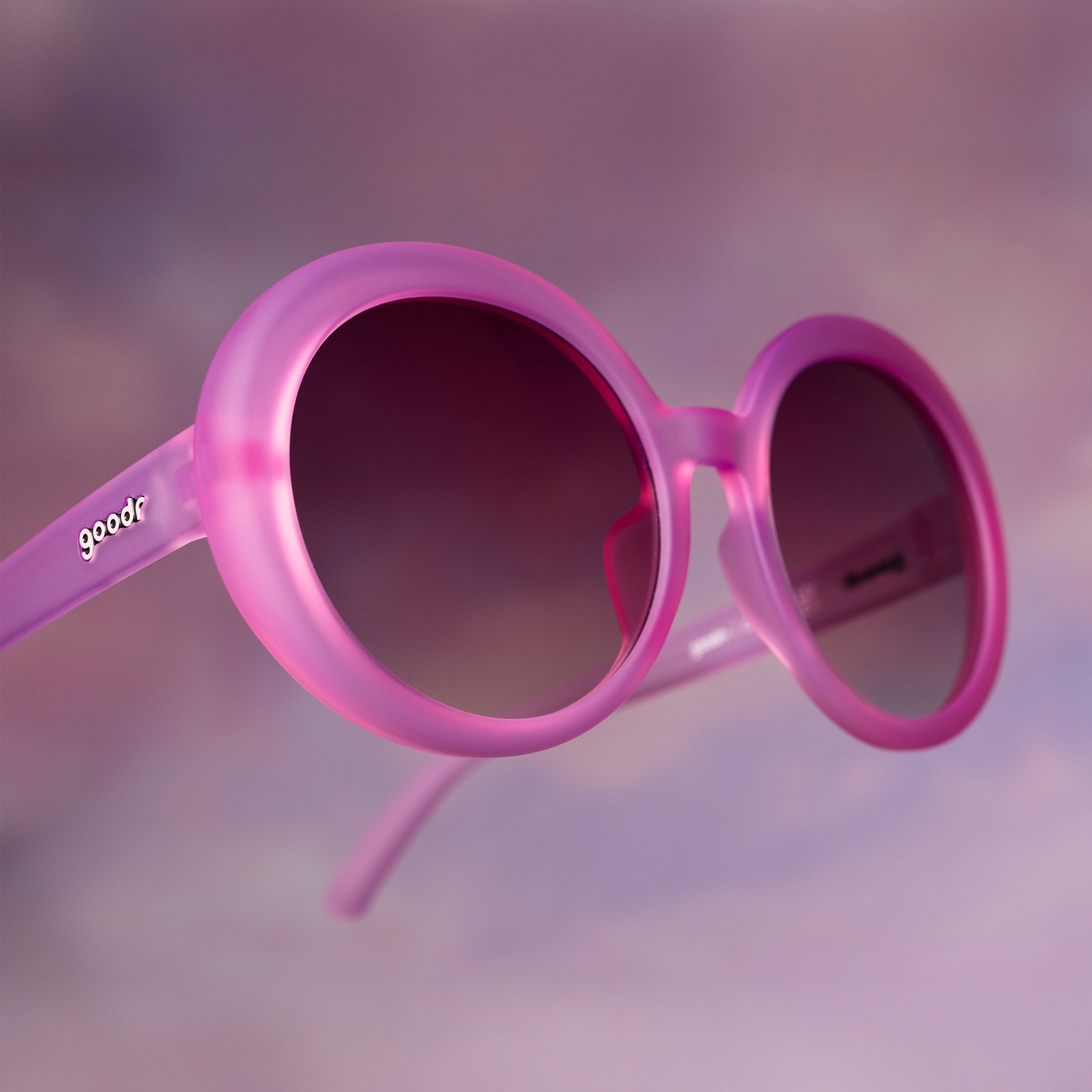 Three-quarter close-up of translucent round pink Mood G sunglasses floating against a purplish cloud-like backdrop.