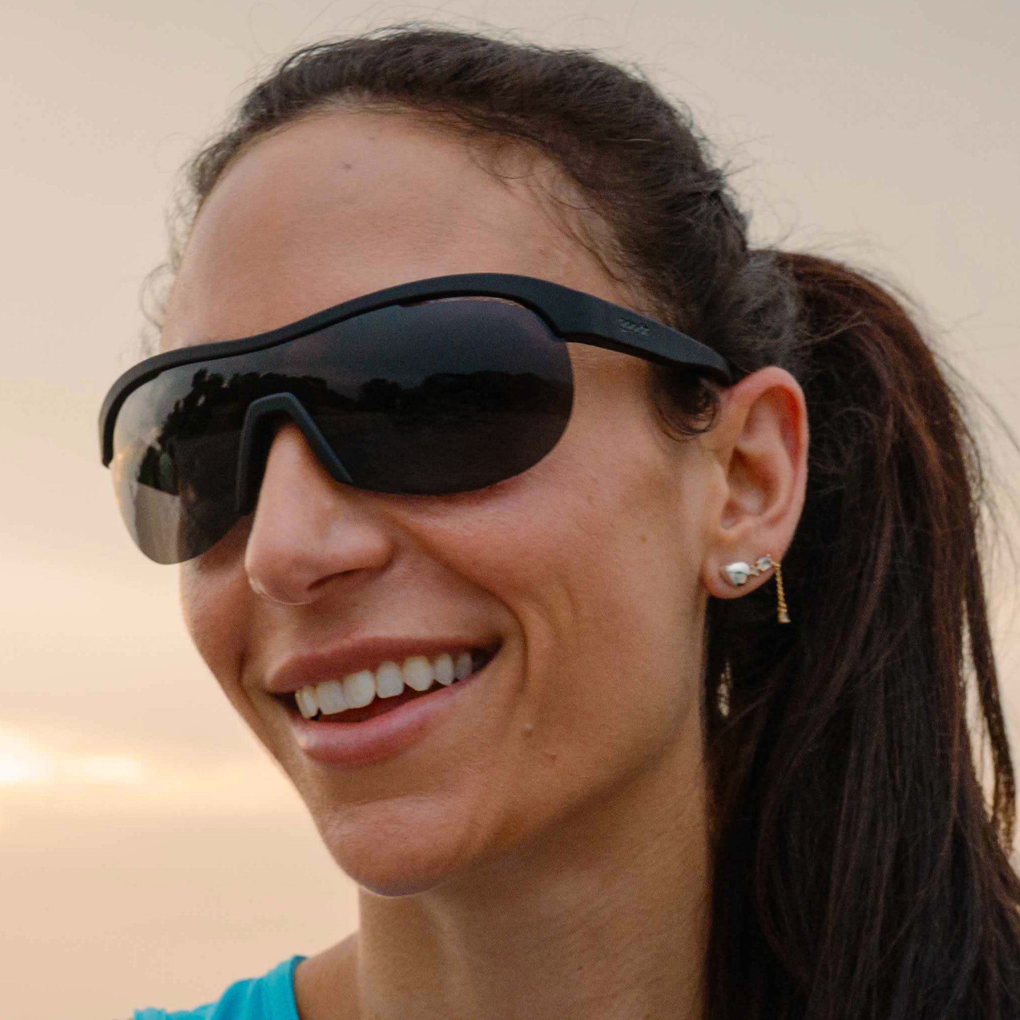 Smiling woman with a ponytail wearing black wraparound shield sunglasses with black polarized lenses.