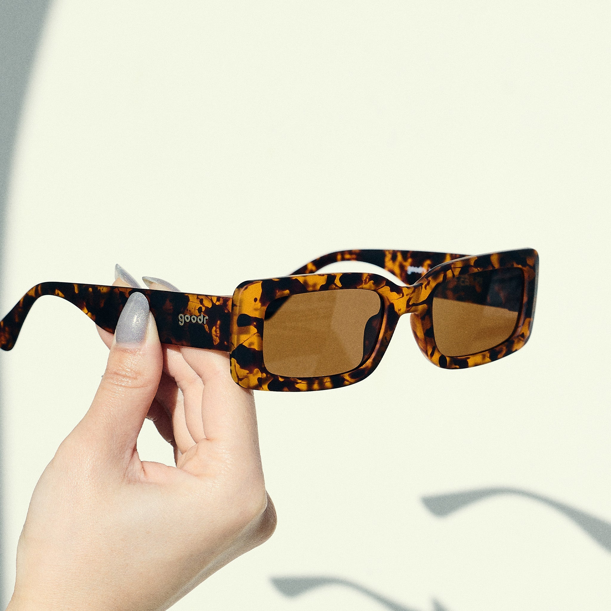 A manicured hand holds up a pair of rectangular sunglasses with brown polarized lenses in front of an off-white backdrop.
