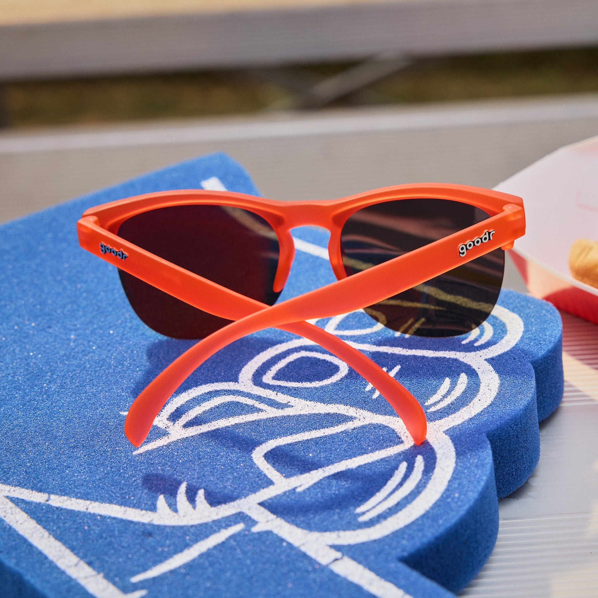 Back angle view of red-orange half-rim sunglasses with orange reflective lenses sitting on top of a blue foam finger on bleachers.