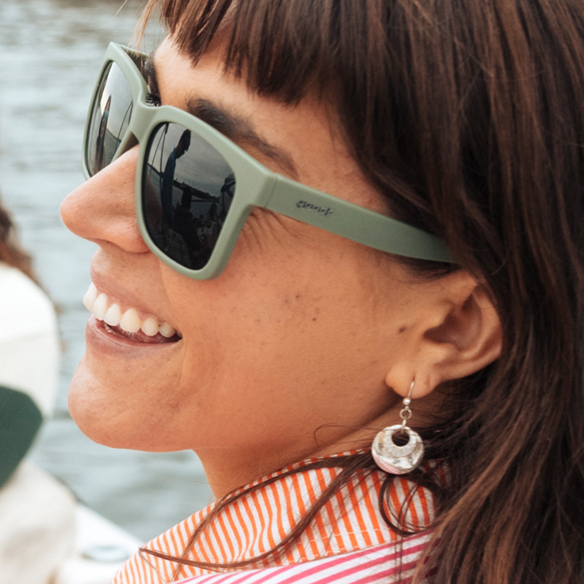 Three-quarter angle view of a laughing young woman with brown hair wearing dark green sunglasses with green lenses.