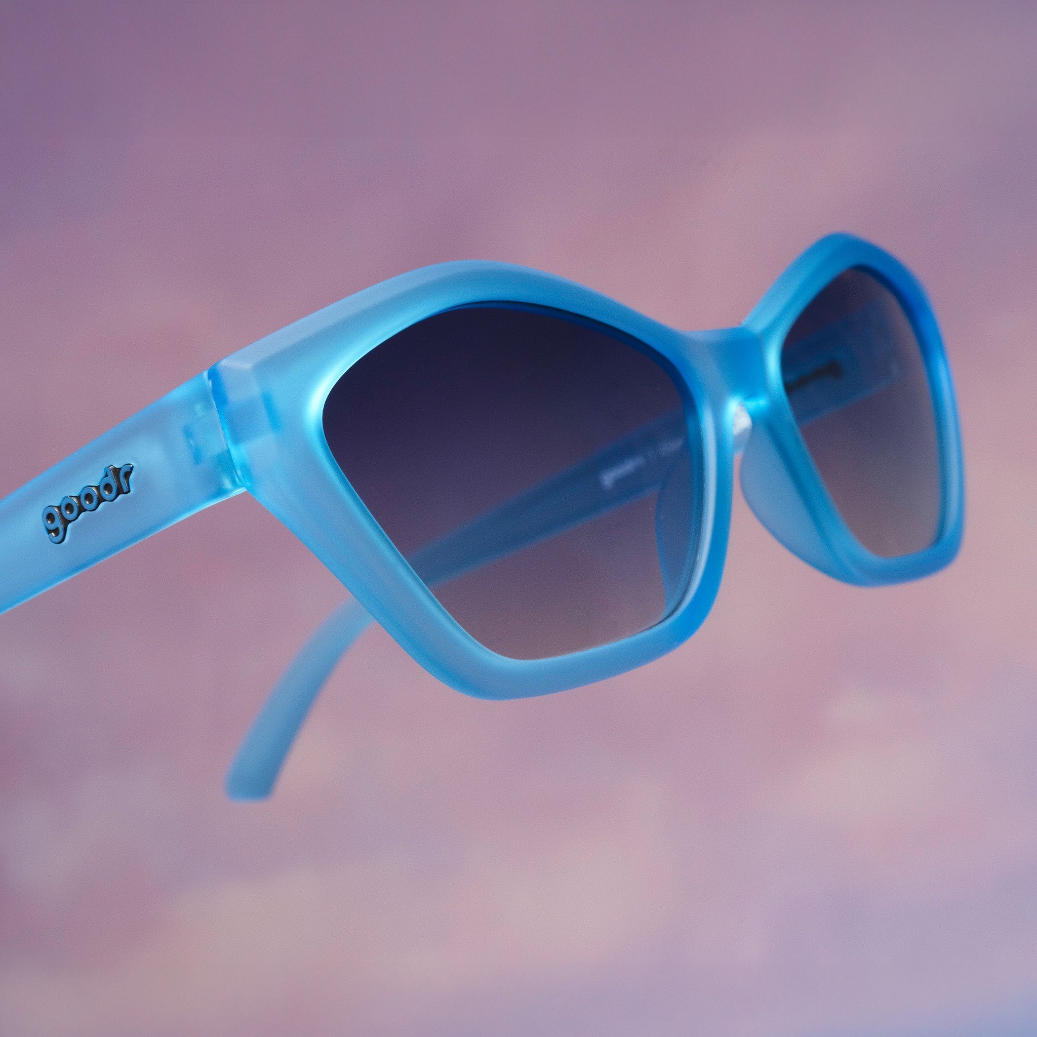 Close-up of a pair of translucent blue geometric cat-eye Deco G sunnies floating against a cloud-like purplish-pink backdrop.