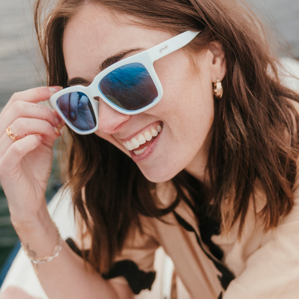 Three-quarter angle view of a laughing young woman with brown hair wearing light blue sunglasses with blue lenses.